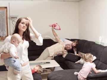 a stressed mother is holding her baby son while her husband and daughters are playing and screaming in the background