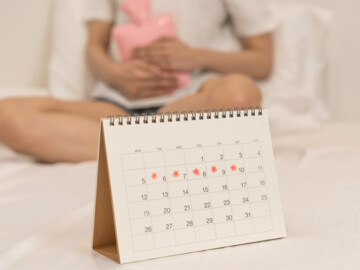 Kalender mit eingezeichneten Menstruationstagen. Im Hintergrund: Frau im Bett mit rosafarbener Wärmflasche auf dem Bauch