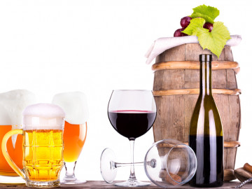 Weintrauben auf einem Weinfass, davor Gläser mit Bier und Wein sowie eine Flasche Rotwein mit Korkenzieher