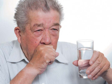 Älterer Mann hustet und hat ein Glas Wasser in der Hand