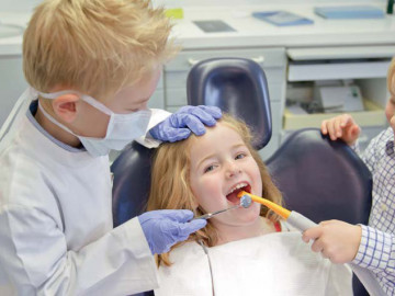 Drei Kinder spielen Zahnarzt. Mädchen sitzt auf dem Behandlungsstuhl, Jungs sind ZFA und ZA